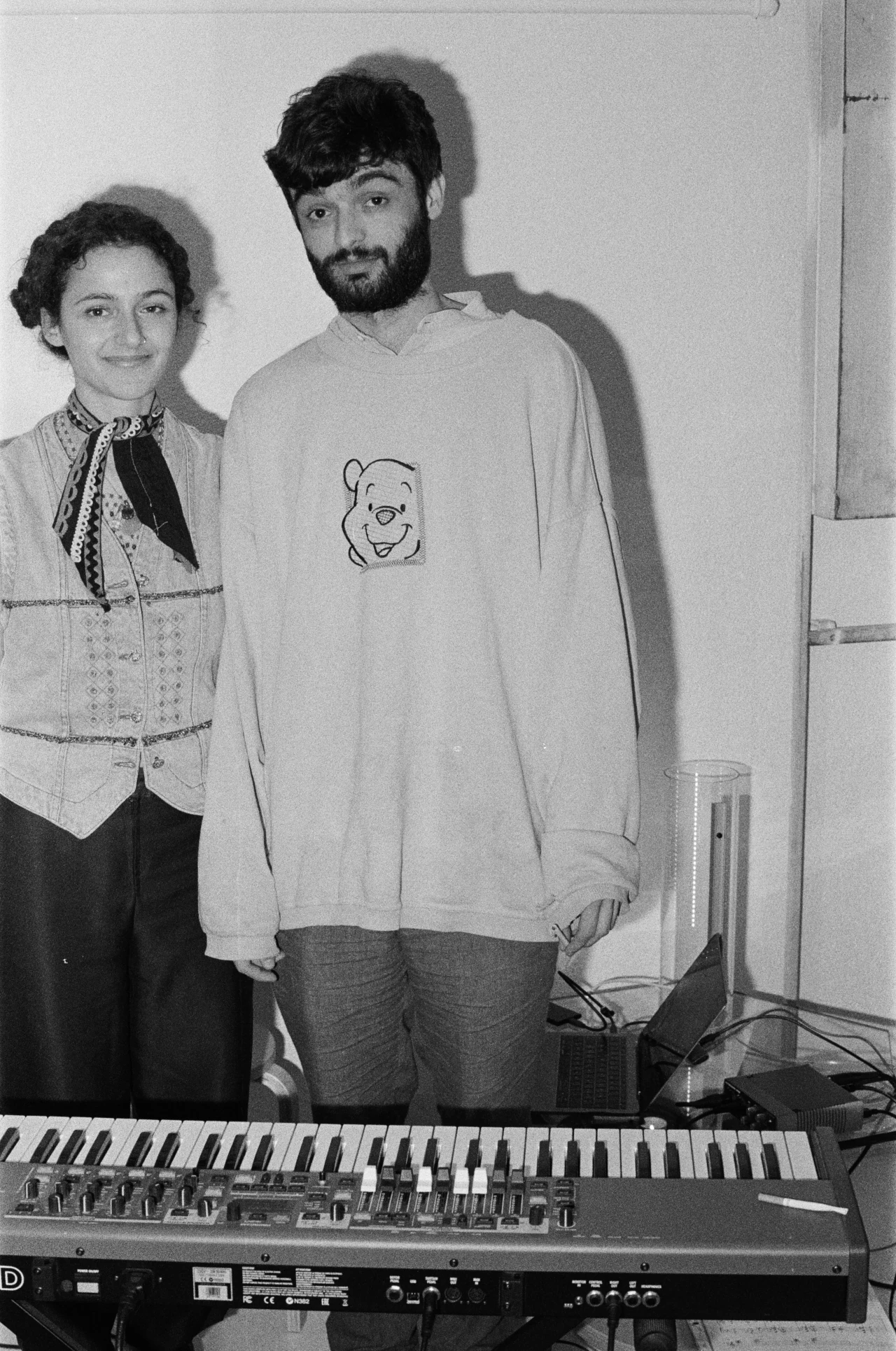 Black and white picture showing Sara and Iannis standing behing a Korg keyboard in a somehow messy room.