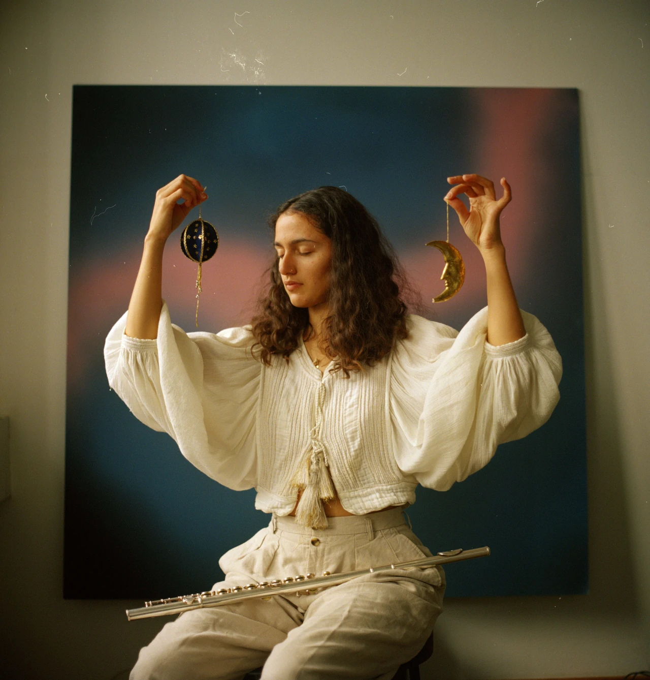 Sara holding up two pendents representing the sun and the moon, the eyes closed, sitting and her flute on the legs